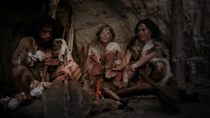 穿着兽皮的史前狩猎采集者部落晚上住在一个山洞里。尼安德特人或智人家庭试图在篝火旁取暖，手握火，做饭