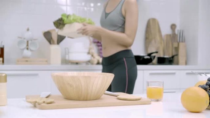 年轻的亚洲女性在厨房喝橙汁制作沙拉，穿着运动服的美丽女性在家中自己使用有机水果和蔬菜制作健康食品。健