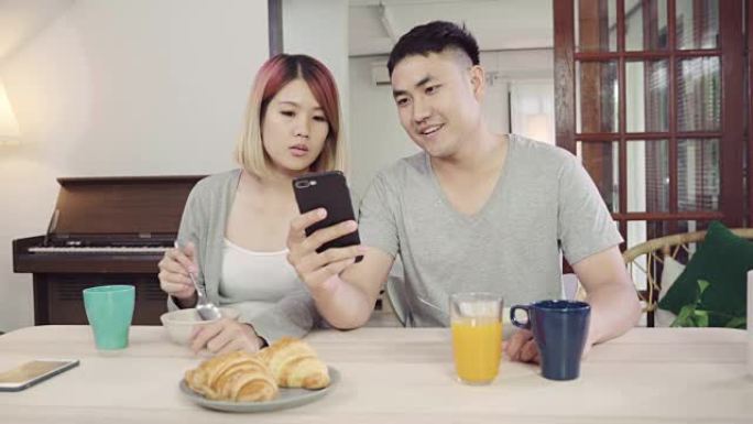 迷人的亚洲年轻夫妇一边吃早餐，一边拿着报纸和手机在餐桌上分心。激动的年轻亚洲夫妇对令人难以置信的好消