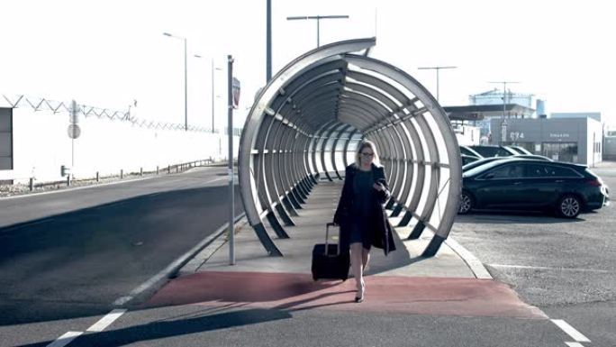 带着智能手机和手提箱的女商人在阳光明媚的机场隧道中行走