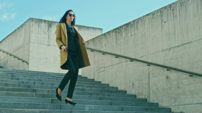 优雅的女商人走下楼梯，使用手机。穿着外套和墨镜的美女在现代城市环境中行走。