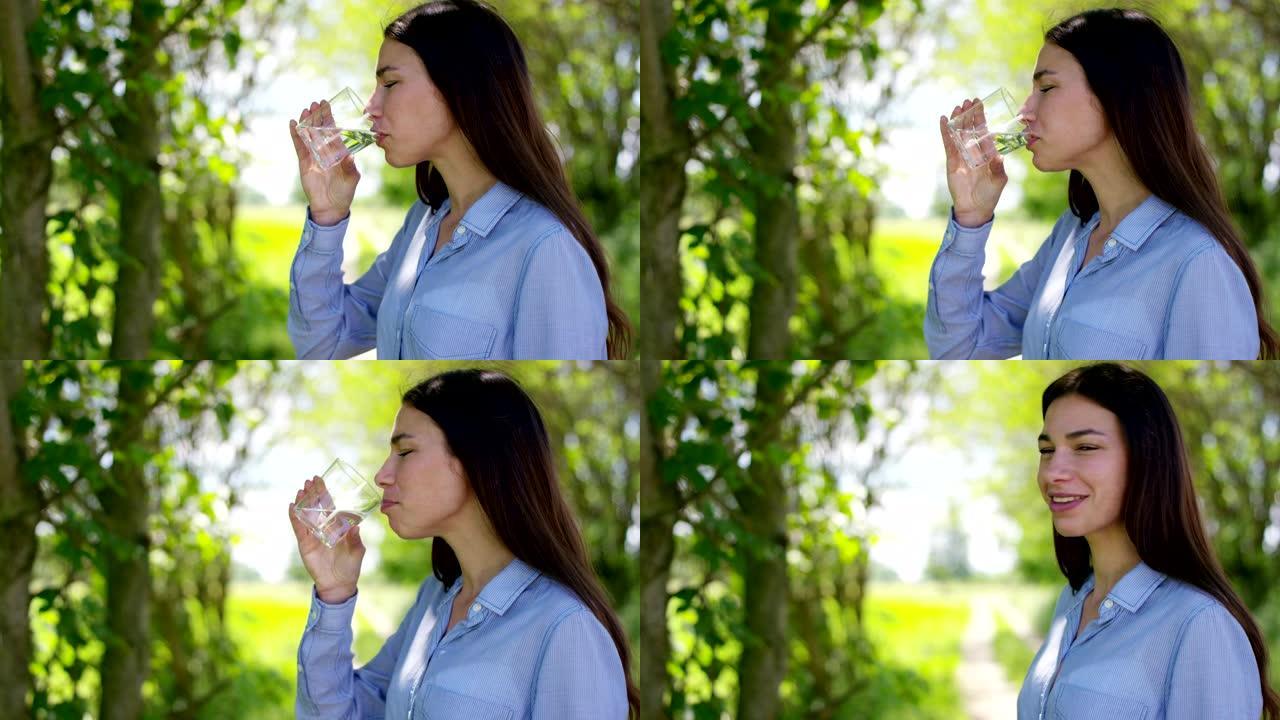 美丽的年轻女孩拿着杯子，喝自然干净的水，在大自然的背景下。