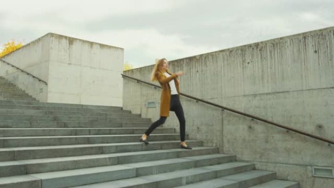 开朗快乐的年轻女子在走下楼梯时积极跳舞。她穿着一件棕色长外套。在商务中心旁边的城市混凝土公园拍摄的场