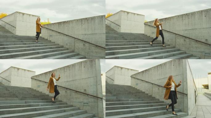 开朗快乐的年轻女子在走下楼梯时积极跳舞。她穿着一件棕色长外套。在商务中心旁边的城市混凝土公园拍摄的场