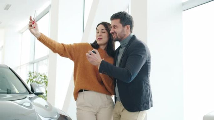 快乐的男人和女人使用智能手机摄像头在经销商处用车钥匙自拍