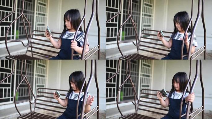 一位亚洲华裔年轻女孩坐在自家后院的秋千上享受下午的时光，同时在手机上玩游戏