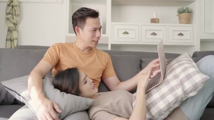 亚洲夫妇在家里的客厅用笔记本电脑搜索网络，甜蜜的夫妇在家放松时躺在沙发上享受爱情时刻。生活方式夫妇在