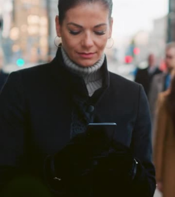 穿着黑色外套的美丽女商人正在市中心的街道上使用智能手机。她走在拥挤的步行街上，看起来很成功。垂直屏幕