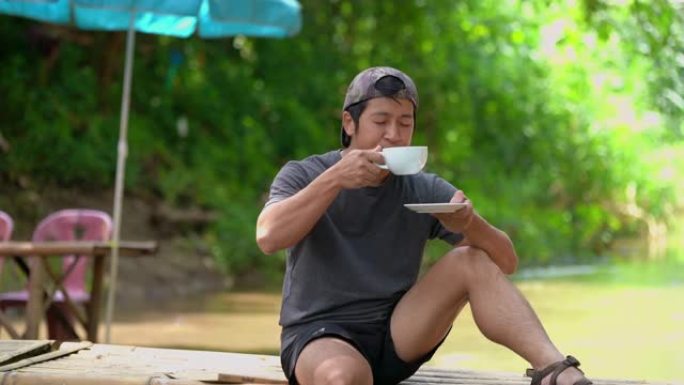 亚洲男子坐在木桥边喝咖啡边用脚玩水
