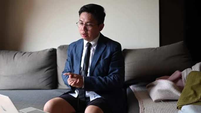 一位打着领带、穿着西装、穿着短裤的亚洲华人中年男子坐在客厅的沙发上，用笔记本电脑与他的商业伙伴和同事