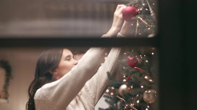 一对年轻夫妇正在他们温暖舒适的家中装饰圣诞树。他们期待这个圣诞节晚上的客人。摄像机穿过房子外面的窗户