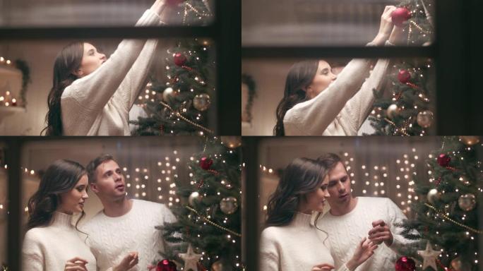 一对年轻夫妇正在他们温暖舒适的家中装饰圣诞树。他们期待这个圣诞节晚上的客人。摄像机穿过房子外面的窗户