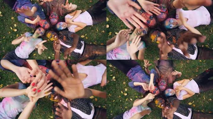 快乐的女孩和男孩躺在公园的草地上，他们的脸和衣服上覆盖着多色油漆，人们在移动手，微笑着看着相机。
