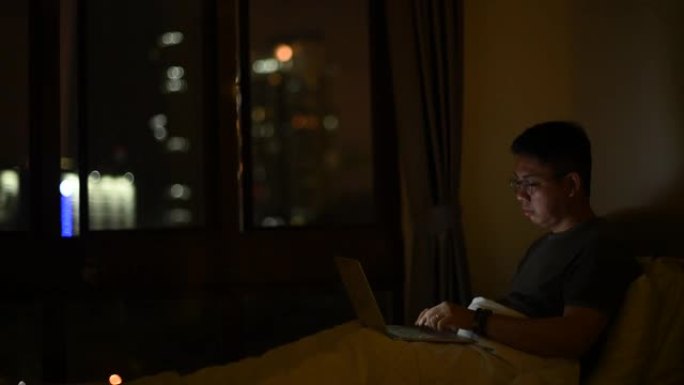 一个亚洲华人中年人晚上在家工作，在床上用他的笔记本电脑