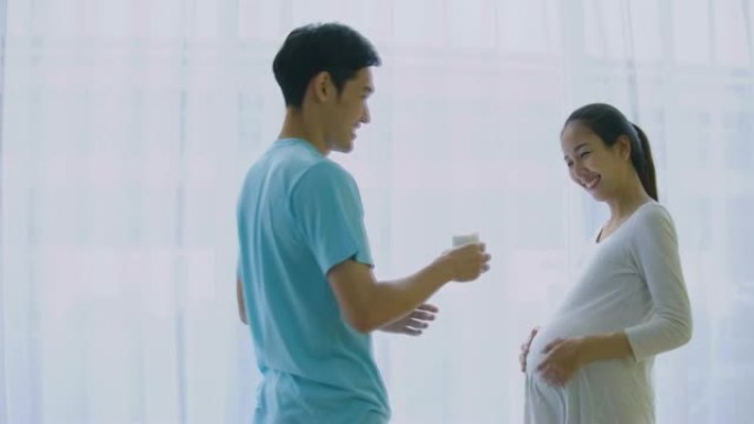 美丽的亚洲孕妇站在客厅里和丈夫一起喝牛奶。