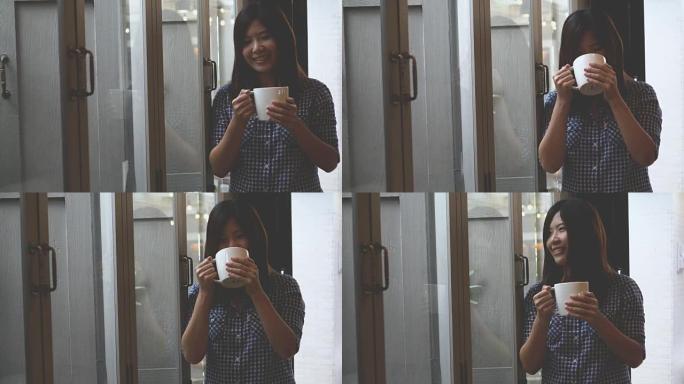 4k镜头亚洲美女站在窗玻璃旁喝着咖啡，带着活泼的微笑，从咖啡馆向外看，生活方式和休闲理念