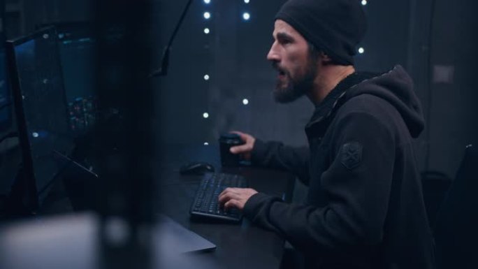 一名留着胡子、戴着帽子、穿着黑色连帽衫的匿名男子正坐在服务器室里操作一台电脑，试图黑进远程服务器。