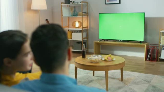 幸福的夫妇坐在家里看绿色色度键屏幕电视，在沙发上放松。情侣房间看体育比赛、新闻、情景喜剧电视节目或电