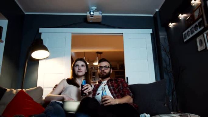快乐的年轻高加索新婚家庭坐在一起在家里看电影，用投影仪慢动作喝酒。