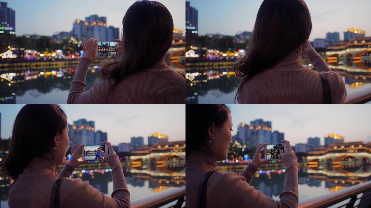 美丽的年轻亚洲女人的侧视图晚上用手机拍摄美丽的城市景色美丽的中国游客在霓虹灯下在河边的九岩桥成都欣赏