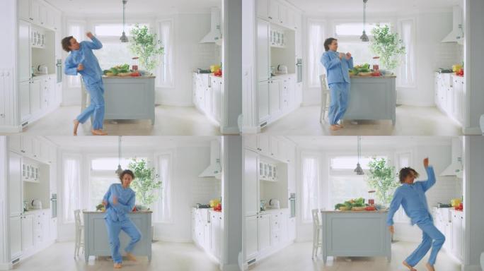 精力充沛的滑稽年轻人，长发穿着蓝色睡衣在厨房跳舞。明亮的白色现代厨房区，桌子上放着健康的绿色蔬菜。舒