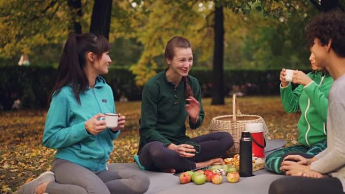 在美丽的秋天，户外瑜伽课结束后，开朗的年轻女性在垫子上休息，野餐聊天和喝茶。可见食物和篮子。