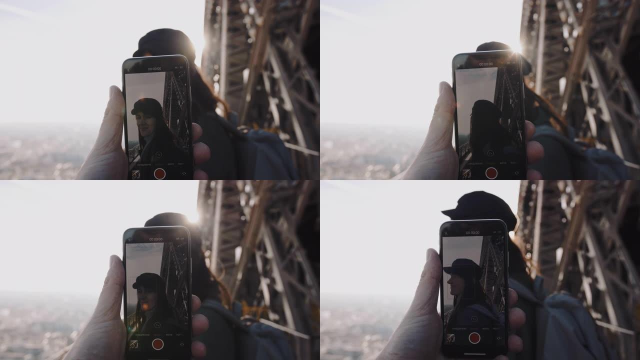 在埃菲尔铁塔上，一名男子手持智能手机拍摄一名开心微笑的女子。浪漫和科技的慢镜头。