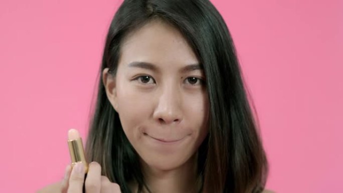 年轻美丽时尚的亚洲女性在粉色背景下用休闲服装化妆刷涂抹嘴唇化妆工作室拍摄。快乐微笑可爱的快乐女人快乐