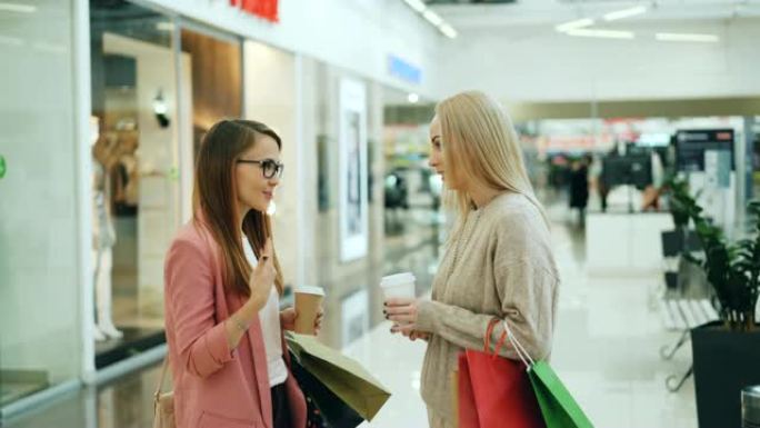 快乐的女同学一起站在商场里，提着袋子和外卖饮料，聊着笑。人体模型上的时尚服装是可见的。