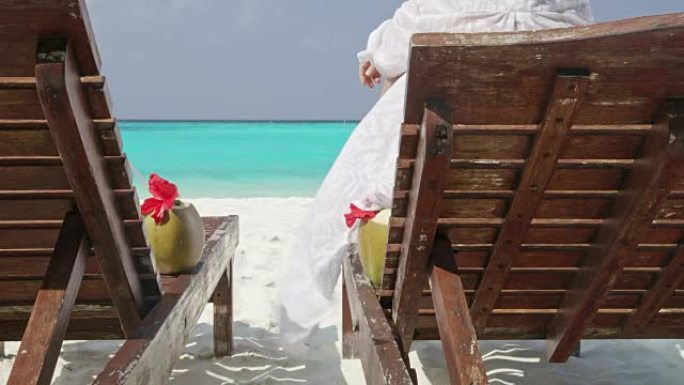 马尔代夫阳光明媚、宁静的热带海洋海滩，女士在躺椅上喝椰子饮料放松