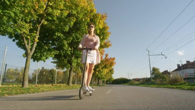 低角度: 年轻女子乘坐电动滑板车沿着秋天的彩色大道通勤。