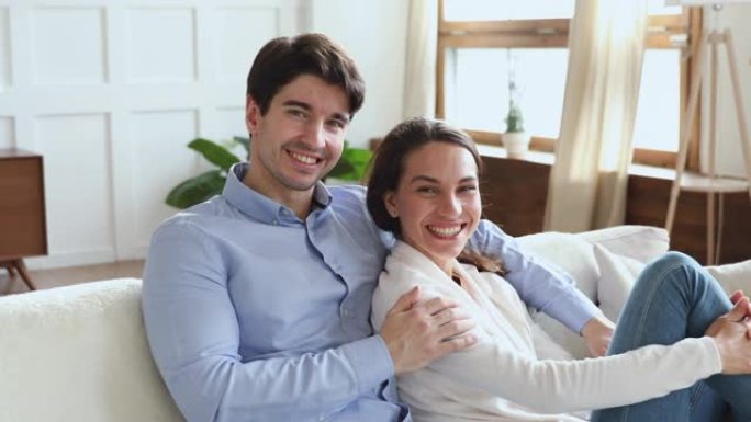 微笑的年轻夫妇拥抱坐在沙发上看着相机