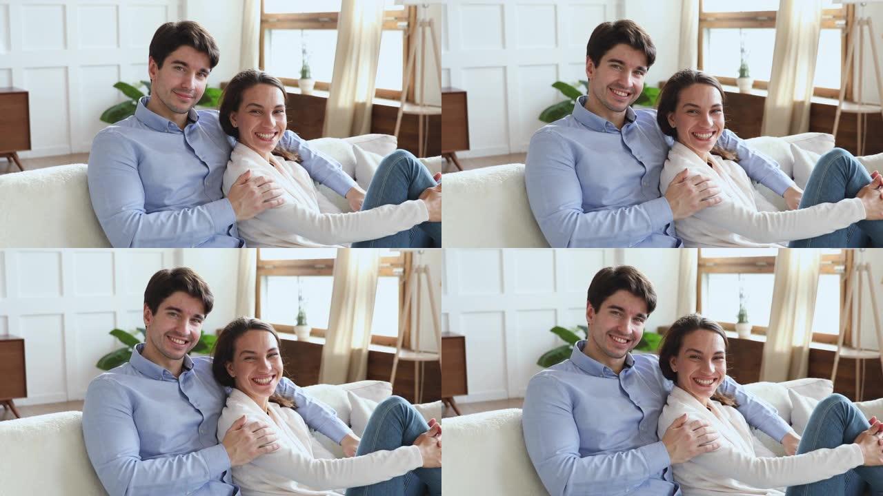 微笑的年轻夫妇拥抱坐在沙发上看着相机