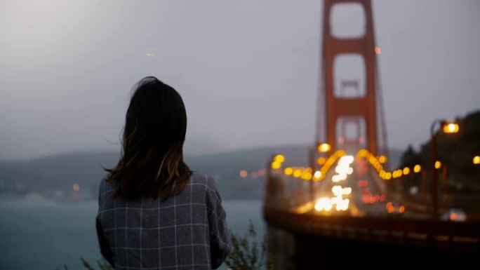 后视图年轻放松的女人在强风的黑暗夜晚观看标志性的金门大桥bokeh交通视图。