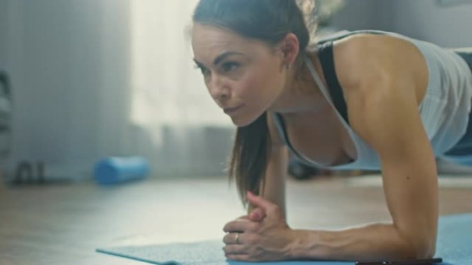 穿着运动运动服的强壮美丽的健身女孩正在用手机上的秒表进行木板运动。她正在室内舒适的客厅里训练。