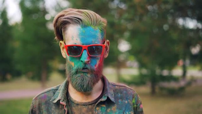 在胡里节上，一个好看的男人站在公园里的肖像，笔直的脸上覆盖着五颜六色的粉末，戴着墨镜看着相机。