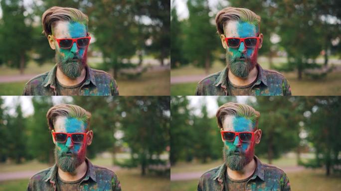 在胡里节上，一个好看的男人站在公园里的肖像，笔直的脸上覆盖着五颜六色的粉末，戴着墨镜看着相机。