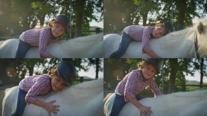 一个戴着骑师头盔的可爱的小女孩的真实照片躺在一匹白色小马马上，在阳光下抚摸着她的马stable