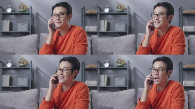 亚洲男子坐在家里客厅的沙发上，享受与朋友在手机上交谈和聊天的乐趣。电信和社会连接技术概念。
