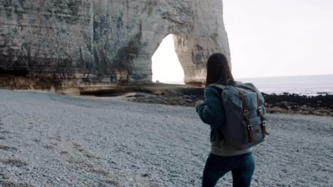 镜头跟随快乐的黑发旅游女人沿着埃特雷塔慢动作附近的海岸走到著名的白色拱形岩石上。
