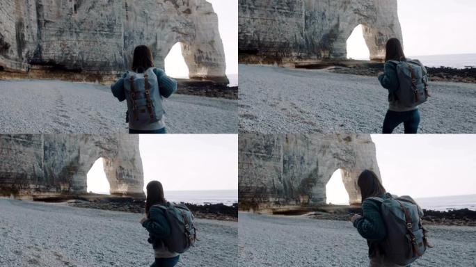 镜头跟随快乐的黑发旅游女人沿着埃特雷塔慢动作附近的海岸走到著名的白色拱形岩石上。
