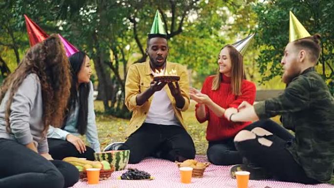 大胡子的非洲裔美国人正在公园举行生日聚会，在蛋糕上吹蜡烛，笑着享受惊喜，他的朋友们鼓掌。