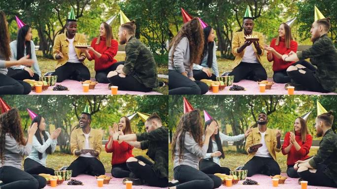 大胡子的非洲裔美国人正在公园举行生日聚会，在蛋糕上吹蜡烛，笑着享受惊喜，他的朋友们鼓掌。
