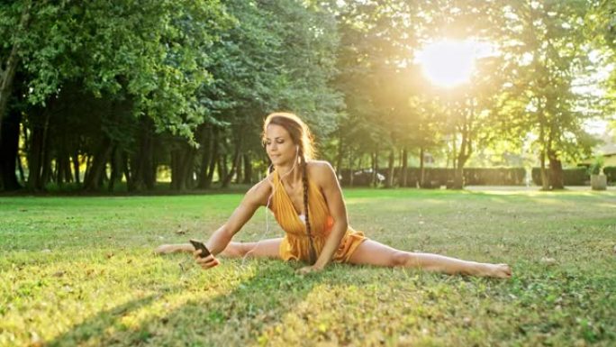 女士灵活的年轻女子练习劈叉，在阳光公园的草地上使用智能手机
