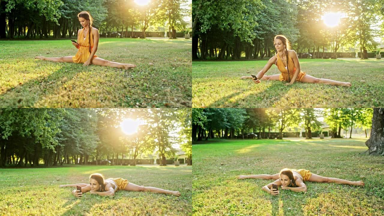 女士灵活的年轻女子练习劈叉，在阳光公园的草地上使用智能手机