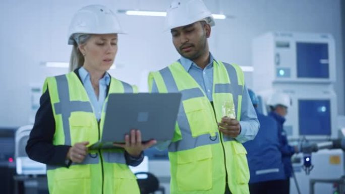 工业设施: 总工程师和项目经理穿着安全背心和安全帽穿过现代工厂，交谈，规划。操作员为生产线编程机械臂
