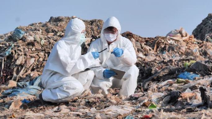 两名持有平板电脑并穿着化学防护服的亚洲科学家在废物病毒COVID-19.Biologists进行研究