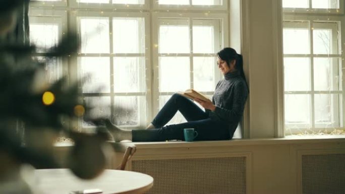 放松的女孩在圣诞节那天看书，坐在窗台上，穿着毛衣和牛仔裤看着窗外的12月。爱好、假期和青年观念。