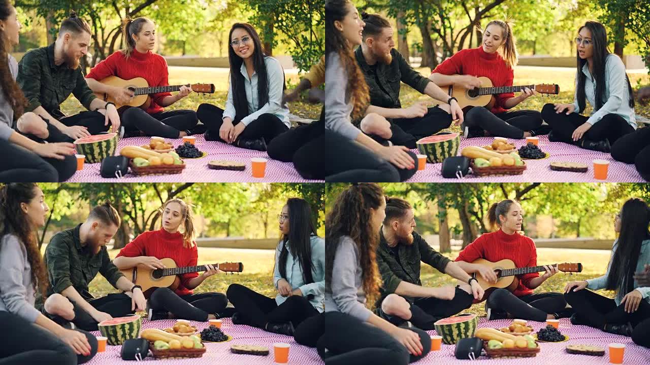 浪漫的年轻人在野餐时唱歌和弹吉他，坐在公园草坪上的格子上，玩得很开心。乐器和文化概念。