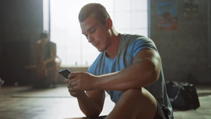 英俊而阳刚的运动青年坐在阁楼健身房的地板上时正在使用智能手机。他在输入信息并思考。他额头上有汗水。妇